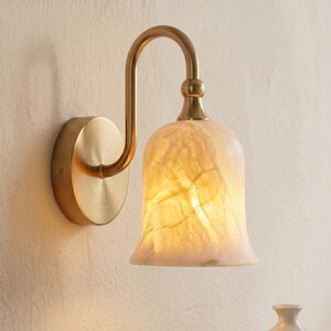 May include: A wall-mounted light fixture featuring a curved, brushed brass arm and a circular wall plate. The bell-shaped lampshade is crafted from translucent, off-white stone with visible natural veining, emitting a warm, soft glow.