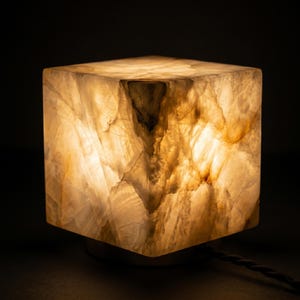 May include: A cube-shaped lamp made of translucent, veined stone that emits a warm, golden glow. The surface features natural, irregular patterns in shades of cream, beige, and amber, with a dark power cord extending from the base.