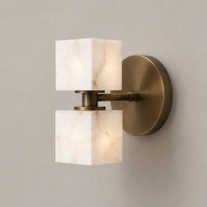 May include: A wall-mounted light fixture featuring two stacked, cube-shaped white alabaster shades with subtle beige veining. The shades are connected by a central metal arm to a circular, brushed antique brass backplate, creating a balanced design.