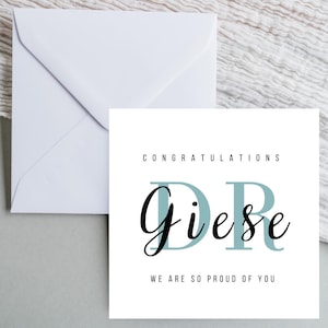 Könnte beinhalten: Weiße Glückwunschkarte mit dem Text "CONGRATULATIONS DR Giese WE ARE SO PROUD OF YOU" in schwarzer und blaugrüner Schrift. Ein weißer Umschlag ist im Hintergrund zu sehen.
