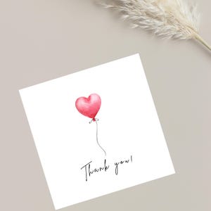 Può includere: Un biglietto bianco con un'illustrazione ad acquerello di un palloncino a forma di cuore rosa. Il palloncino è attaccato a un sottile filo grigio. Le parole "Thank you!" sono scritte in un delicato script sotto.