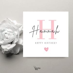 Può includere: Biglietto di auguri bianco con il nome "Hannah" in corsivo nero e una grande "H" rosa. Sotto, le parole "HAPPY BIRTHDAY" e un piccolo cuore rosa. Un fiore bianco è visibile a sinistra.