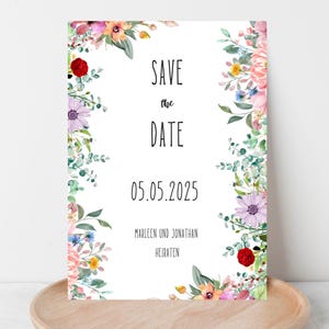 Könnte beinhalten: Eine florale "Save the Date" Karte mit weißem Hintergrund. Die Karte zeigt bunte Aquarellblumen und Grün, die den Text "SAVE the DATE" und das Datum "05.05.2025" umrahmen. Die Namen "Marleen und Jonathan" sind ebenfalls enthalten.