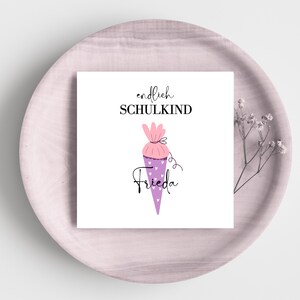 Puede incluir: Una tarjeta cuadrada blanca con las palabras "endlich SCHULKIND" y el nombre "Frieda" en escritura. La tarjeta presenta un diseño de cono escolar morado y rosa con acentos de corazón. La tarjeta está sobre un plato rosa claro.