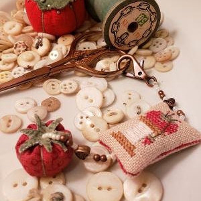 May include: A small cross-stitch pillow with a red and white design of a sewing machine and a spool of thread. The pillow is attached to a beaded chain with a lobster clasp. There are two red fabric pincushions shaped like strawberries, a spool of thread, and a pair of scissors on a white surface.
