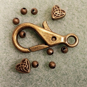 May include: A gold-toned metal clasp with a heart-shaped charm featuring a Celtic knot design. The clasp is surrounded by small round beads.