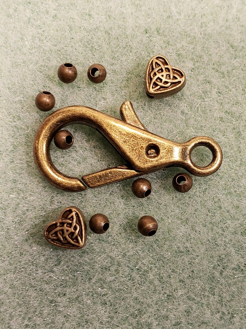 May include: A gold-toned metal clasp with a heart-shaped charm featuring a Celtic knot design. The clasp is surrounded by small round beads.