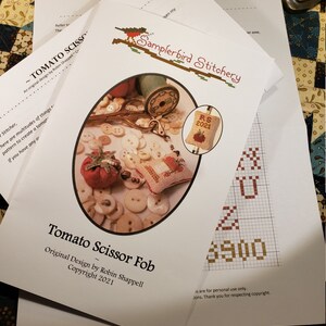 May include: A white paper pattern for a cross stitch project called "Tomato Scissor Fob". The pattern includes a small tomato, scissors, and buttons. The text "RS 2021" is visible in the image.
