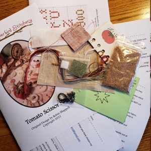 May include: A cross stitch kit for a tomato scissor design. The kit includes fabric, floss, beads, and a pattern. The pattern is titled "Tomato Scissor" and includes the text "Original Design by Robin Shappell Copyright 2021".
