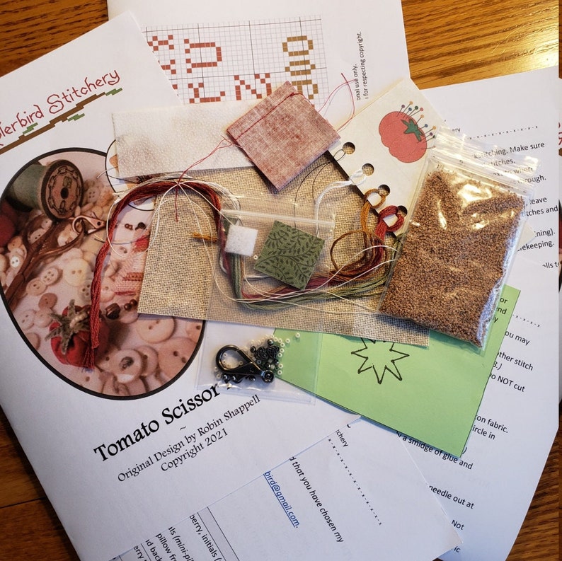 May include: A cross stitch kit for a tomato scissor design. The kit includes fabric, floss, beads, and a pattern. The pattern is titled "Tomato Scissor" and includes the text "Original Design by Robin Shappell Copyright 2021".