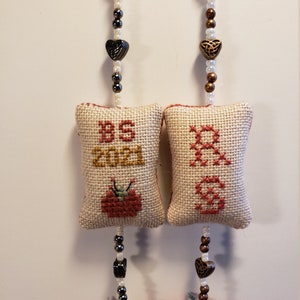 May include: Two handmade fabric ornaments with a red and green tomato design. Each ornament has a white fabric pillow with cross-stitch embroidery. One pillow says "BS 2021" and the other says "Grandma". Each ornament has a beaded chain with a heart charm.
