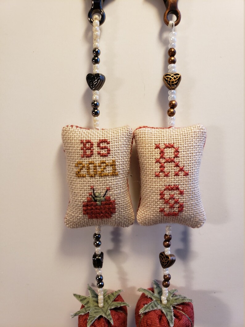 May include: Two handmade fabric ornaments with a red and green tomato design. Each ornament has a white fabric pillow with cross-stitch embroidery. One pillow says "BS 2021" and the other says "Grandma". Each ornament has a beaded chain with a heart charm.