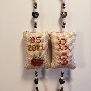 May include: Two handmade fabric ornaments with a red and green tomato design. Each ornament has a white fabric pillow with cross-stitch embroidery. One pillow says "BS 2021" and the other says "Grandma". Each ornament has a beaded chain with a heart charm.