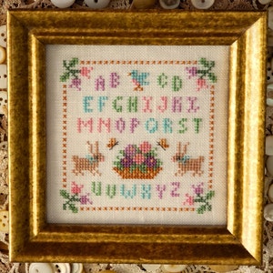 May include: A framed cross-stitch sampler featuring the alphabet in colourful letters, two bunnies, and a basket of flowers. The letters are in a light pink, blue, and green colour scheme. The bunnies are brown and white. The basket is brown with a pink and purple floral design.