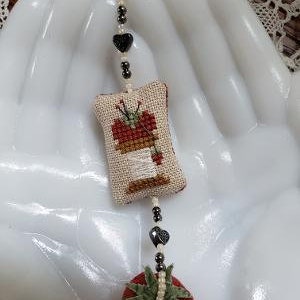 May include: A white fabric pincushion with a cross-stitch design of a spool of thread and a needle. The pincushion is attached to a beaded chain with a red fabric tomato charm.