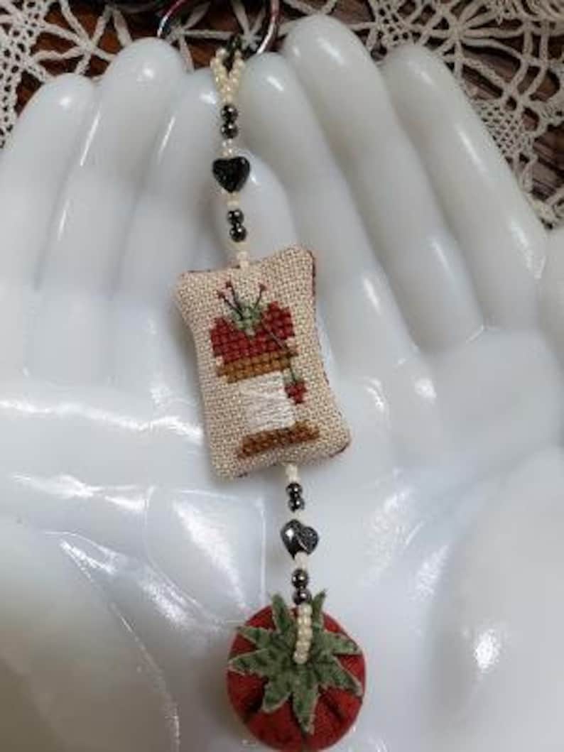 May include: A white fabric pincushion with a cross-stitch design of a spool of thread and a needle. The pincushion is attached to a beaded chain with a red fabric tomato charm.