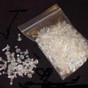 May include: A clear plastic bag filled with small, clear plastic push pins. The bag is open and some of the push pins are scattered on a black surface.