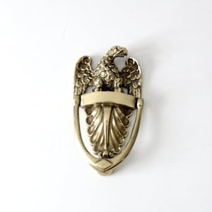 May include: A brass door knocker shaped like an eagle with its wings spread. The eagle is perched on a decorative base with a blank rectangular plate.