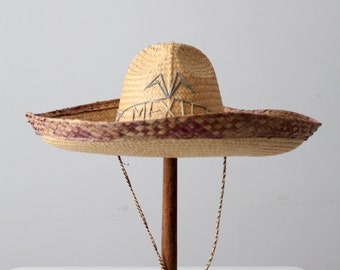 May include: A straw sombrero with a red and brown trim and a woven design. The hat has a brown cord with red beads hanging from it.