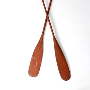 May include: Two wooden oars with red paint, crossed in an X shape against a white background.