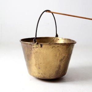 Puede incluir: Un cubo de latón vintage con un asa de alambre y un poste de transporte de madera. El cubo tiene una pátina desgastada y envejecida y es perfecto para la decoración rústica o el estilo de granja.