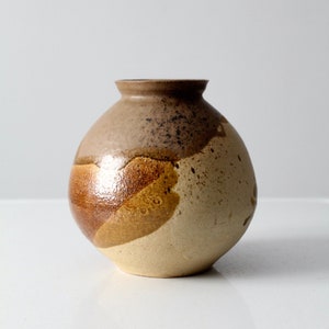 May include: A round ceramic vase with a narrow neck and a wide, spherical body. The surface features a multi-toned glaze with earthy shades of beige, tan, and dark brown, accented by speckled textures and smooth, glossy patches across the exterior.