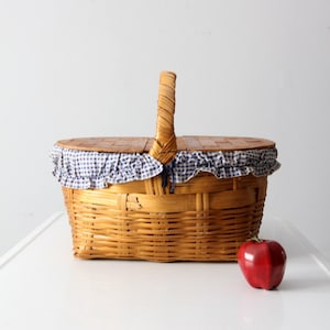 May include: A vintage wicker picnic basket with a blue and white checkered lining and a wooden handle. A red apple sits in front of the basket.