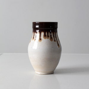 Puede incluir: Un jarrón de cerámica vintage con cuerpo blanco y borde marrón oscuro. El esmalte marrón gotea por los lados, creando un patrón único. El jarrón tiene forma redondeada y una abertura ligeramente ensanchada.