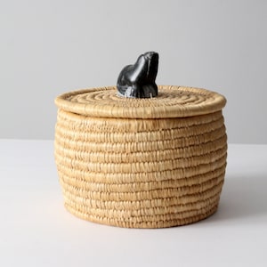 Może przedstawiać: Okrągły, wiklinowy kosz z pokrywką ozdobioną rzeźbą morskiego ssaka. Kosz wykonany jest z naturalnego słomy i ma jasnobrązowy kolor.