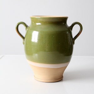 Puede incluir: Un jarrón de cerámica verde con dos asas y un borde blanco. El jarrón tiene un cuerpo redondeado y un labio ligeramente acampanado.