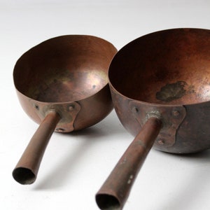 May include: Two copper ladles with long handles. The ladles are slightly different sizes and have a worn, aged look.