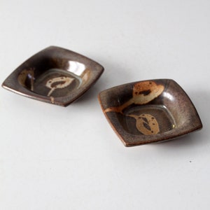 Può includere: Due piccole ciotole quadrate in ceramica con smalto marrone e beige. Le ciotole hanno un bordo leggermente rialzato e una superficie testurizzata.