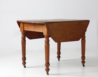 May include: A vintage wooden drop leaf table with turned legs and casters. The table is made of solid wood and has a natural finish. It is in good condition with some signs of age and use.