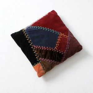 Könnte beinhalten: Ein Patchwork-Kissen aus Samtstoff in den Farben Rot, Schwarz, Blau, Braun und Orange. Das Kissen ist mit buntem Garn zusammengenäht.