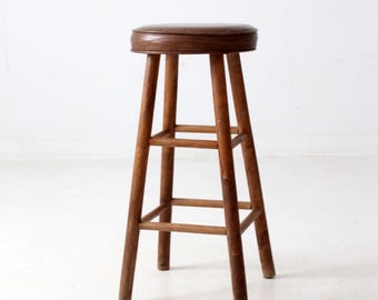 May include: A vintage wooden bar stool with a round, brown leather seat. The stool has four wooden legs connected by horizontal supports. The wood has a warm, natural finish. The stool is a classic piece of furniture.
