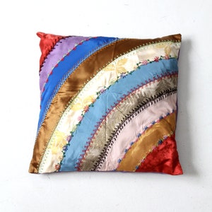 Könnte beinhalten: Ein quadratisches Kissen mit Patchwork-Muster. Das Kissen besteht aus verschiedenen Stoffen in den Farben Rot, Blau, Lila und Gold. Die Stoffe sind in einem geschwungenen Muster zusammengenäht, wodurch ein Regenbogeneffekt entsteht.