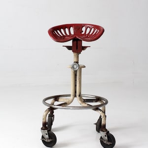 May include: A rolling stool featuring a red metal tractor-style seat with slotted cutouts. The base is constructed from off-white metal tubing with a circular footrest and four black caster wheels. A central adjustment knob is visible on the stem.