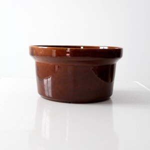 May include: A vintage, oval-shaped, brown ceramic casserole dish. The dish has a glossy finish and a slightly flared rim. The interior is a darker shade of brown than the exterior. The dish is likely used for baking and serving.