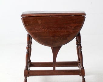 May include: A vintage, dark brown, drop-leaf table. The table has a semi-circular top that folds down, supported by turned legs and a central support. The table is approximately 30 inches tall.