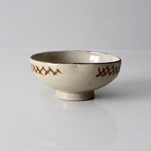 Può includere: Una piccola ciotola in ceramica rotonda con smalto color crema e base a piedistallo. L'esterno presenta un motivo a tratteggio incrociato marrone dipinto a mano che circonda il bordo superiore, mentre l'interno e il bordo sono di colore beige.