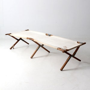 May include: A white canvas cot with wooden legs and metal hardware. The cot is folded up and ready to be used.