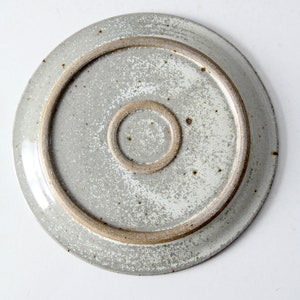 May include: The underside of a round, white ceramic plate with a speckled glaze. The plate has a brown rim and a smaller brown circle in the center.