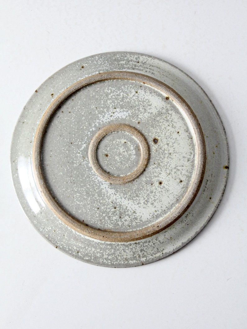 May include: The underside of a round, white ceramic plate with a speckled glaze. The plate has a brown rim and a smaller brown circle in the center.