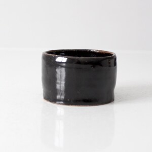 Peut inclure: Un petit gobelet cylindrique en céramique doté d'une glaçure noire profonde et brillante. Le récipient présente une forme façonnée à la main, légèrement irrégulière, avec un bord supérieur brun foncé visible, offrant un design simple.