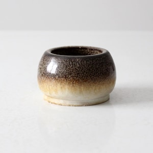 May include: A small, round ceramic bowl with a speckled brown and tan glaze. The bowl has a wide opening and a slightly flared base. The glaze transitions from dark brown at the top to tan and white at the base. The bowl is likely vintage.