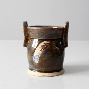 Peut inclure: Un petit vase en céramique brun avec deux anses. Le vase a un glaçage brun marbré avec des accents bleus et blancs.