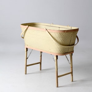 May include: A vintage wicker baby bassinet with a light pink trim and four wooden legs. The bassinet is made of woven wicker and has a rectangular shape.