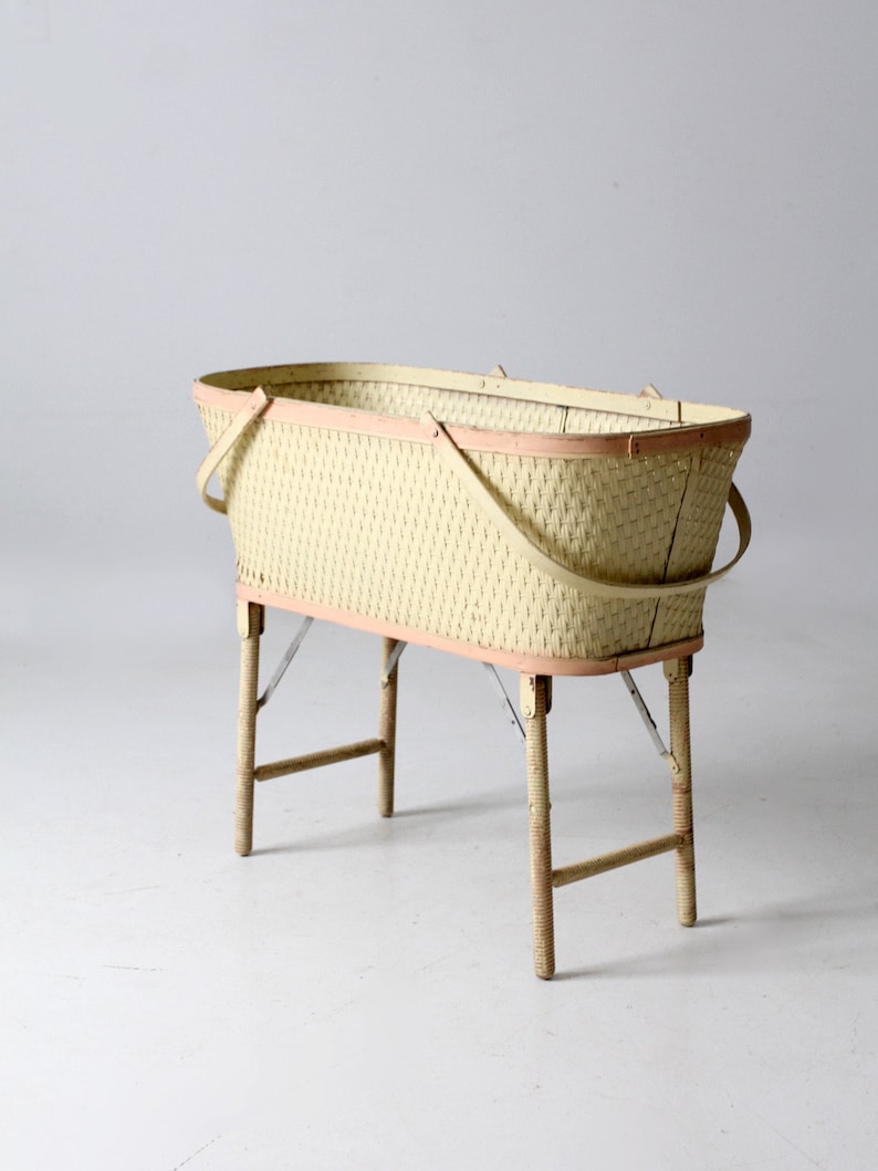 May include: A vintage wicker baby bassinet with a light pink trim and four wooden legs. The bassinet is made of woven wicker and has a rectangular shape.