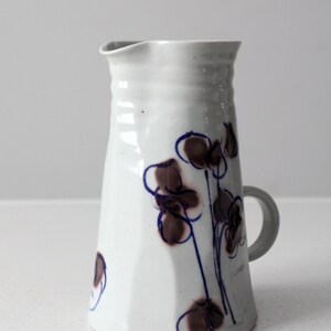 May include: White ceramic pitcher with a brown and blue floral design. The pitcher has a handle and a wide mouth.