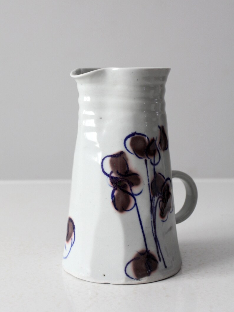 May include: White ceramic pitcher with a brown and blue floral design. The pitcher has a handle and a wide mouth.